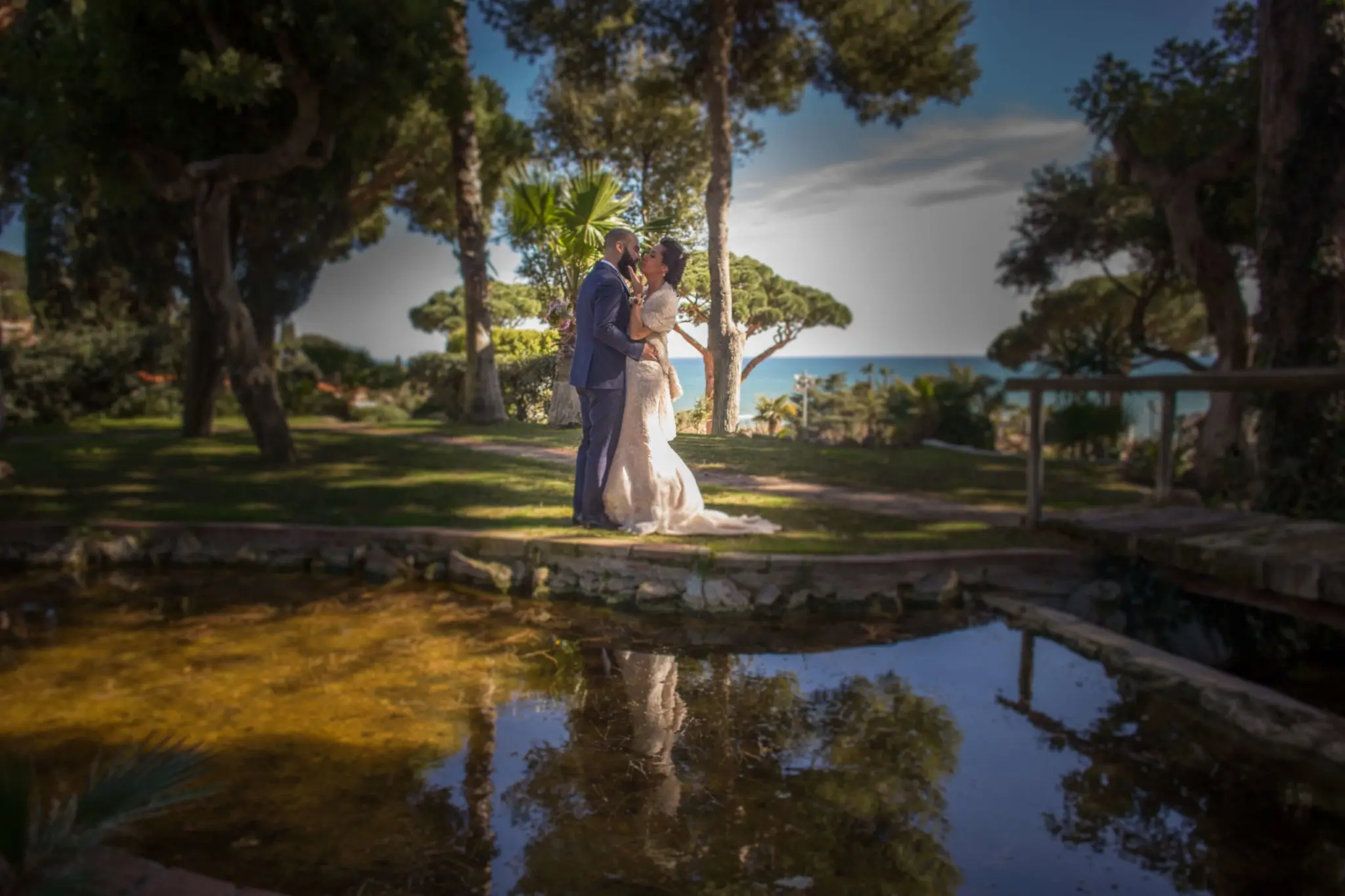 Una pareja se besa junto a un estanque, entre pinos y con el mar al fondo, a la luz baja de la tarde